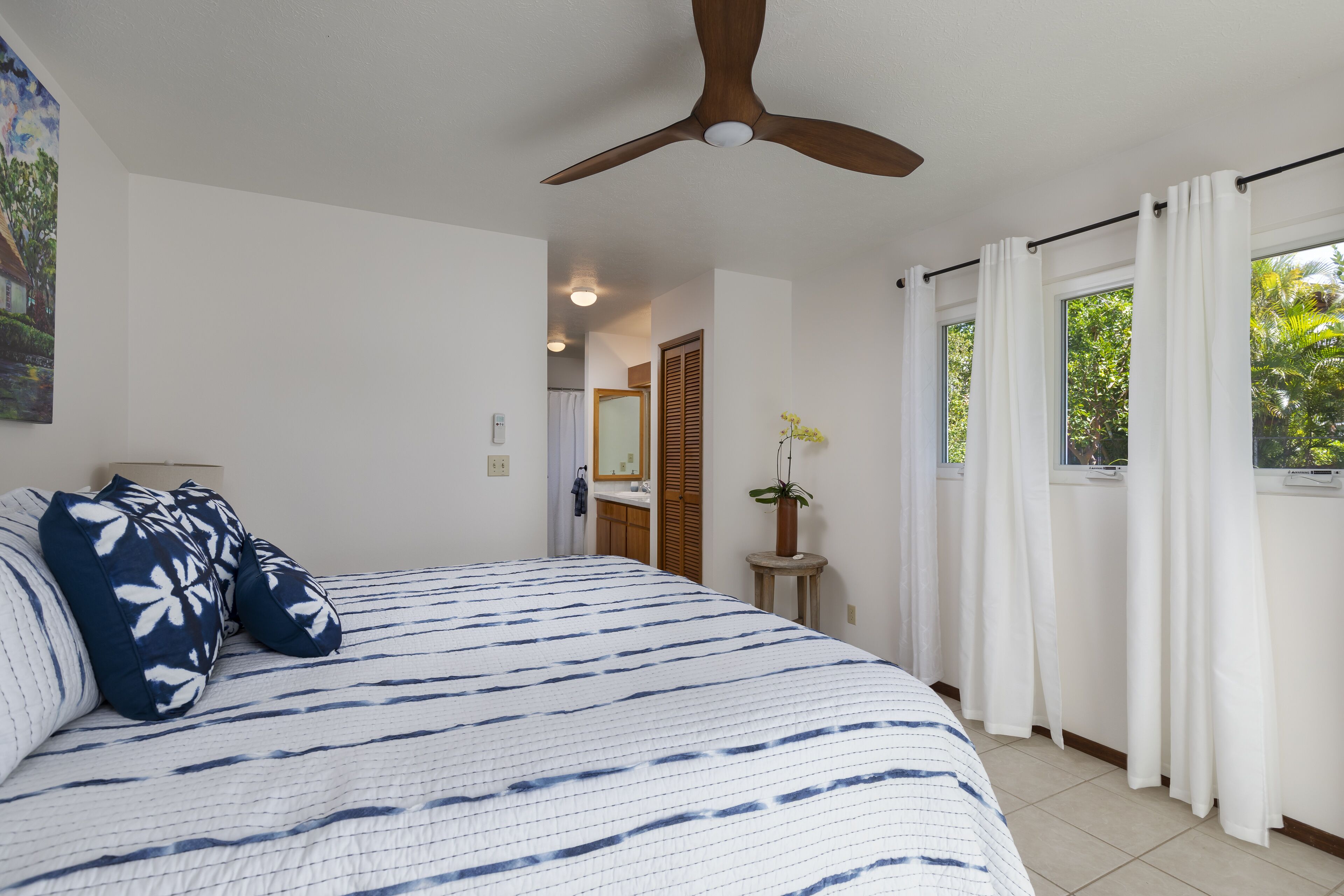 Une chambre à coucher avec un grand lit, un ventilateur de plafond, une fenêtre avec des rideaux et une petite table sur laquelle est posée une plante.