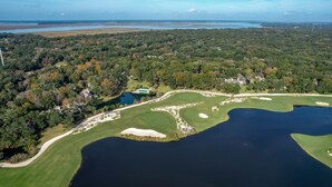 Luchtbeeld van een golfbaan met meerdere bunkers, een vijver en omliggende beboste gebieden.