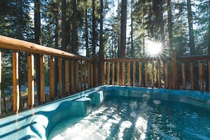 Un jacuzzi rodeado por una terraza de madera y altos árboles, con luz solar filtrándose.