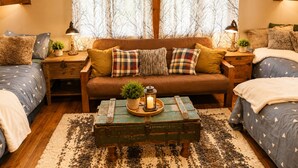 A cozy bedroom with a wooden ceiling, a brown sofa, two bedside tables with lamps, a green trunk table, and a large window with sheer curtains. - Private Hot Tub in the Pines in North Lake Tahoe (Carnelian Bay)