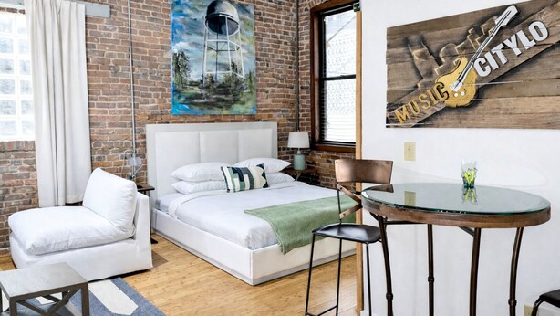 Una habitación de hotel moderna con pared de ladrillo, una cama grande, una zona de estar con una silla y una mesita, y obras de arte en las paredes.