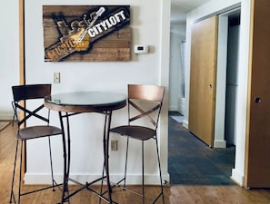 A dining area with a round table and two chairs, a wall-mounted sign with a guitar and the word "CITYLOFT", and a hallway leading to a bathroom.