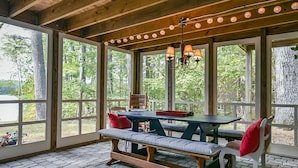 A covered outdoor dining area with a wooden table and benches, surrounded by large windows offering a view of trees and a body of water.