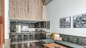 Eine moderne Küche mit dunklen Holzausziehtüren, Edelstahlgeräten und einer Bar mit Barhockern. Im Wohnbereich befindet sich eine grüne Couch und ein Holztisch, an der Wand hängen zwei Schwarz-Weiß-Fotos.