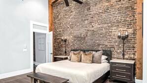 Ein Schlafzimmer mit Backsteinwand, einem Bett mit vielen Kissen, einer Bank, zwei Nachttischen und einem Deckenventilator.