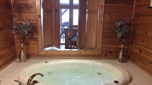 Ein Badezimmer einer Holzhütte mit einer großen Badewanne, zwei Vasen mit Trockenblumen und einem Fenster mit Blick auf eine Holzterrasse und Stühle.