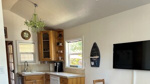 A kitchen with wooden cabinets, a dishwasher, a dining table with chairs, a chandelier, and a television mounted on the wall. - Cozy Private Beach Bungalow, New Hot Tub,  1 Dog ok, 3 short Blocks To Beach. (Santa Cruz)
