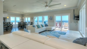 A spacious living room with a large white sofa, a dining area with a table and chairs, and a balcony with a view of the ocean.