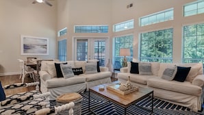 A spacious living room with two white sofas, a coffee table, and a large window with blinds. - Sandestin Beachside 4BR | Golf Cart + Pool (Miramar Beach)