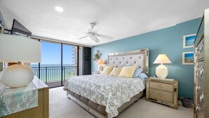 A bedroom with a large bed, two bedside lamps, a nightstand, a dresser, and a view of the ocean through a balcony.