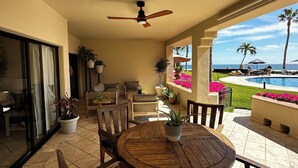 Um alpendre coberto com uma mesa redonda de madeira e cadeiras, plantas em vasos, vista para uma piscina e oceano.