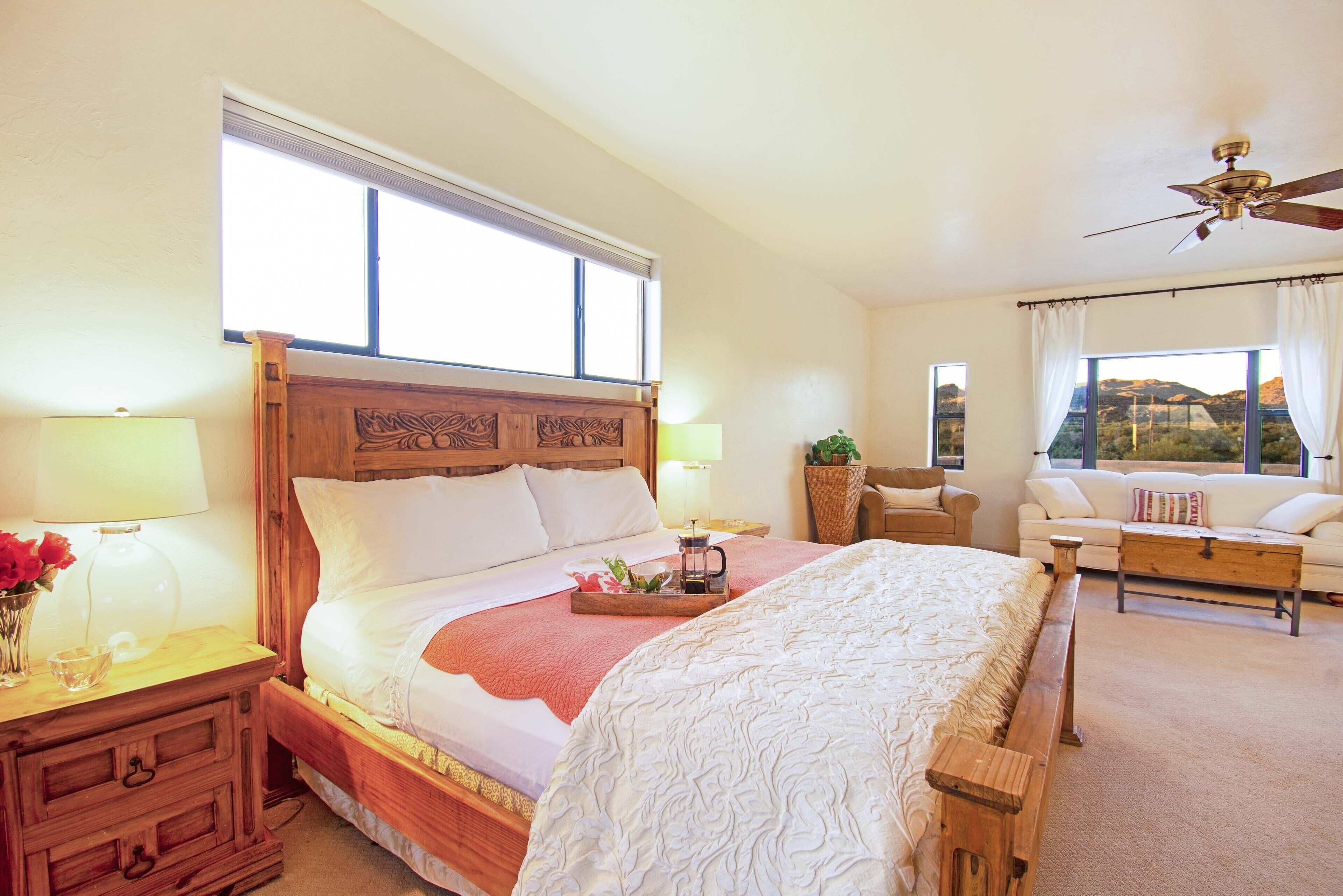 A spacious bedroom with a large bed, a wooden headboard, a nightstand, a bedside table, a lamp, a window with curtains, a ceiling fan, a sofa, and a view of the outdoors.