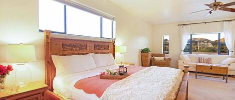 A spacious bedroom with a large bed, a wooden headboard, a nightstand, a bedside table, a lamp, a window with curtains, a ceiling fan, a sofa, and a view of the outdoors.