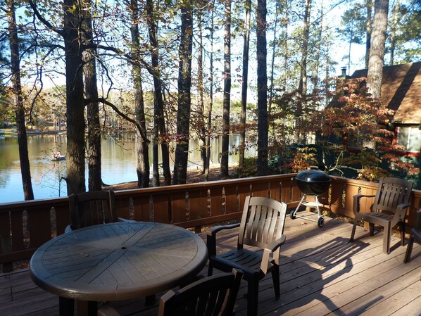 A wooden deck with a round table and two chairs, overlooking a lake with boats and surrounded by trees.