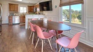 A kitchen with wooden cabinets, a stainless steel refrigerator, and a dining area with a wooden table and pink chairs. - Experience family fun at Turtle Time, a 4-bedroom home in Oak Island, NC. (Oak Island)