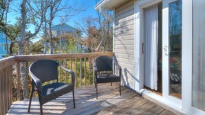 A wooden deck with two wicker chairs, a sliding glass door, and a view of trees and a house in the background. - Experience family fun at Turtle Time, a 4-bedroom home in Oak Island, NC. (Oak Island)