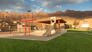 Un parque infantil con tobogán y columpios, rodeado por una zona vallada, con edificios y una montaña al fondo.