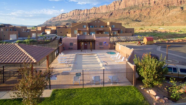 Un hotel con zona de piscina, aparcamiento vallado y montañas al fondo.