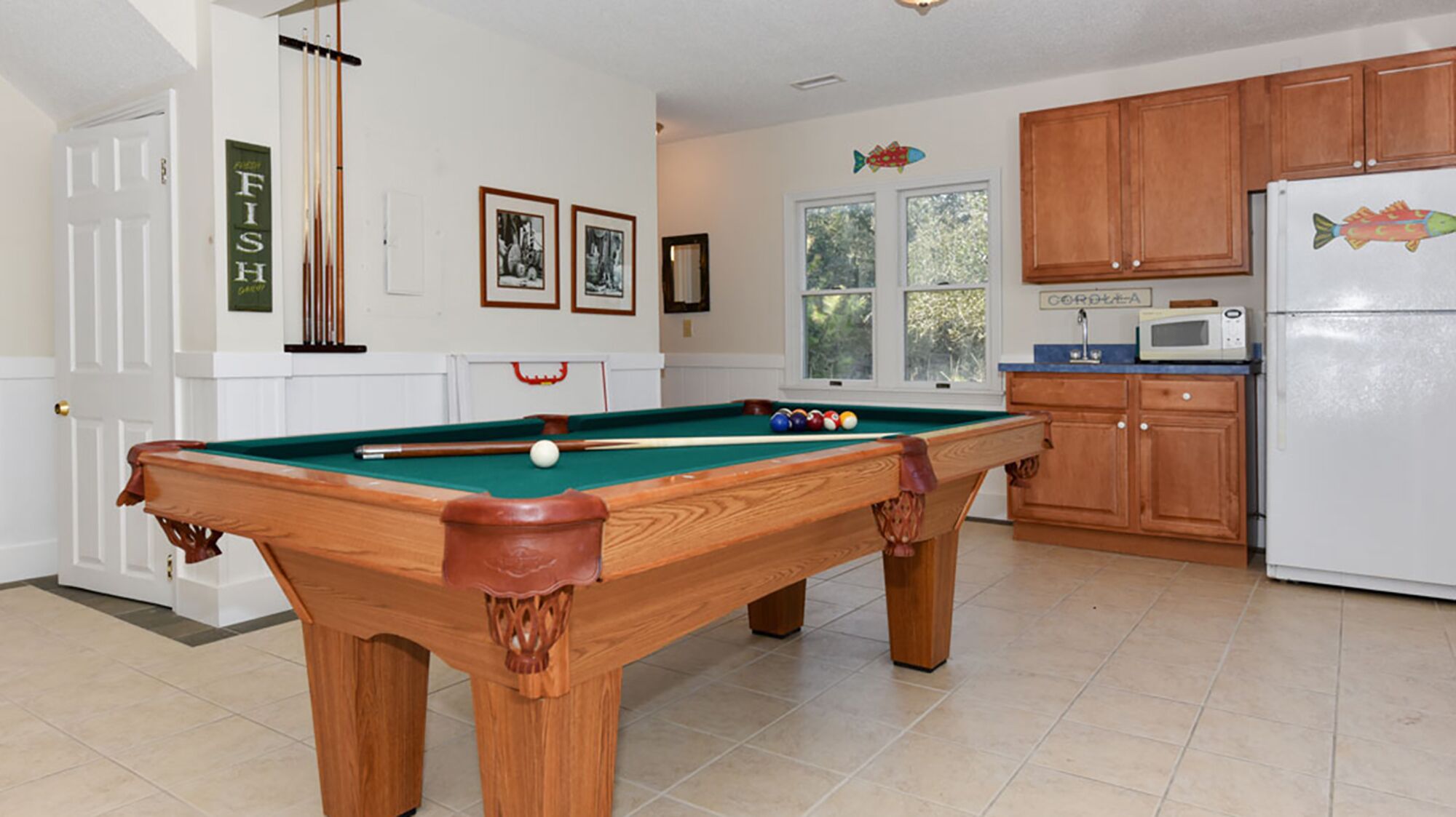 A pool table in a room with a kitchen area, wooden cabinets, and framed pictures on the wall.