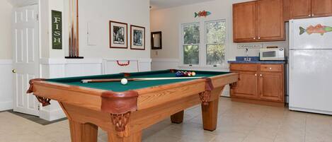A pool table in a room with a kitchen area, wooden cabinets, and framed pictures on the wall.