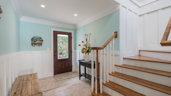 Un couloir avec un escalier en bois, une porte donnant sur l’extérieur, une petite table sur laquelle est posé un vase de fleurs et un élément décoratif mural.