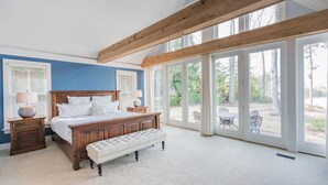 A bedroom with a large bed, wooden headboard, and a bench. There are two bedside tables with lamps. The room has large windows and a view of the outdoors.