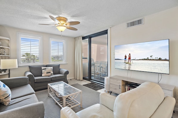 A modern living room with a large flat-screen TV, grey sofas, a glass coffee table, and a ceiling fan.
