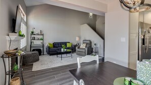 Un salon moderne avec une table à manger en bois foncé, un canapé noir agrémenté de coussins verts et un escalier menant à un espace cuisine.