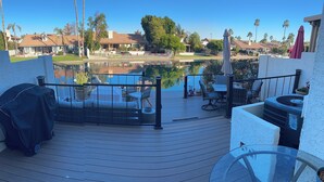 Une terrasse en bois avec des meubles d’extérieur, une table en verre et une vue sur un lac et des maisons au loin.