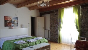 Une chambre avec un lit, une table de chevet blanche, une couverture verte, un plancher en bois, un mur en pierre et une fenêtre avec des rideaux verts.