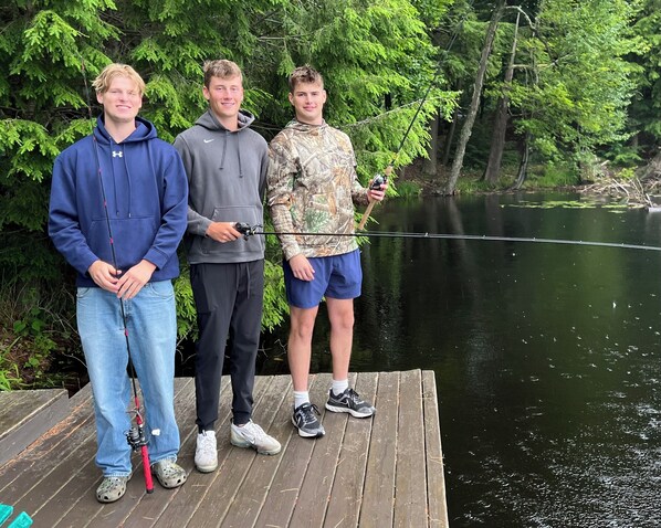 Tres personas de pie en un muelle de madera junto a un lago, con vegetación densa al fondo.