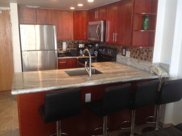 A modern kitchen with wooden cabinets, stainless steel appliances, a marble countertop, and a bar area with stools.