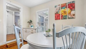 Un coin repas avec une table ronde blanche, des chaises blanches, une plante en pot et une fenêtre avec des stores.