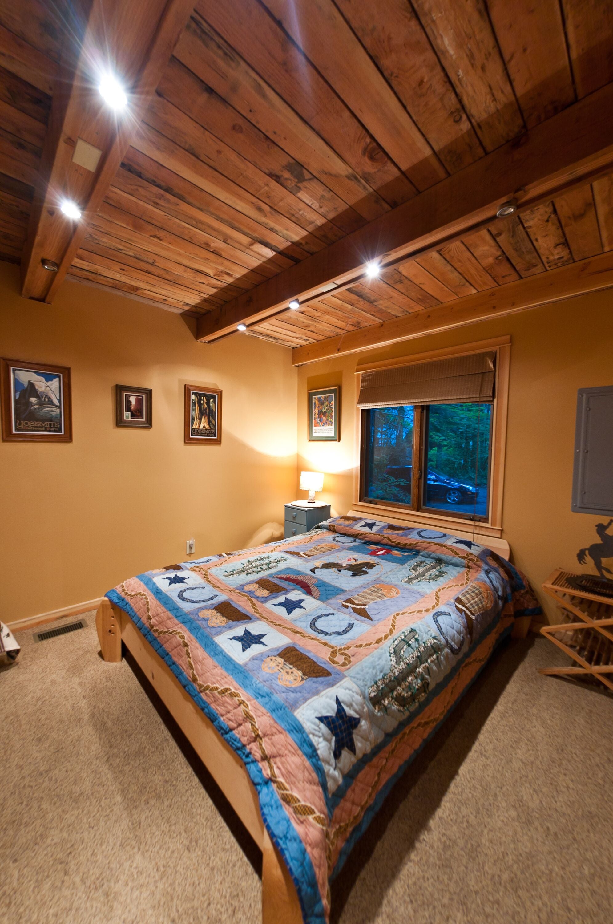 Ein Schlafzimmer mit Holzbalkendecke, einem großen Bett mit Bettdecke, einem Nachttisch, einer Lampe und einem Fenster mit Blick auf Bäume.