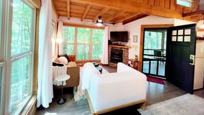 Interior acogedor de una cabaña con chimenea, sofá, mesa de comedor y ventanas grandes que dan a un bosque.