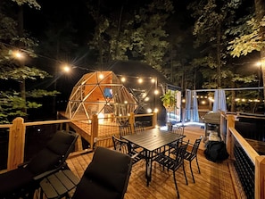A wooden deck with a dining table and chairs, surrounded by trees and a geodesic dome structure. - The Hummingbird Treehouse (Monteagle)
