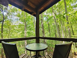 A covered outdoor seating area with a table and chairs, surrounded by a wooden railing, overlooking a forested area. - The Hummingbird Treehouse (Monteagle)