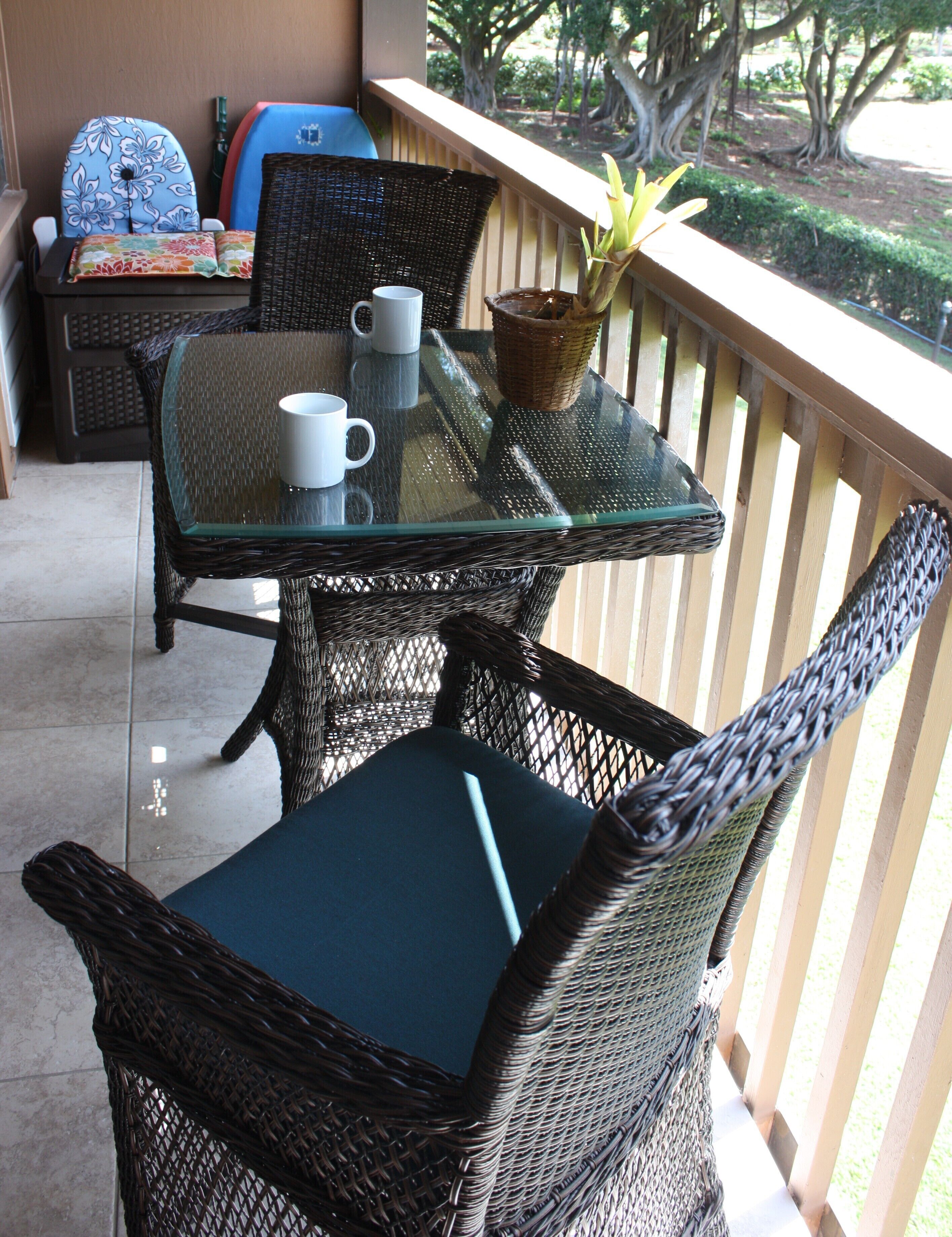 A glass-top table with two white mugs and a woven basket on a balcony with wicker chairs.