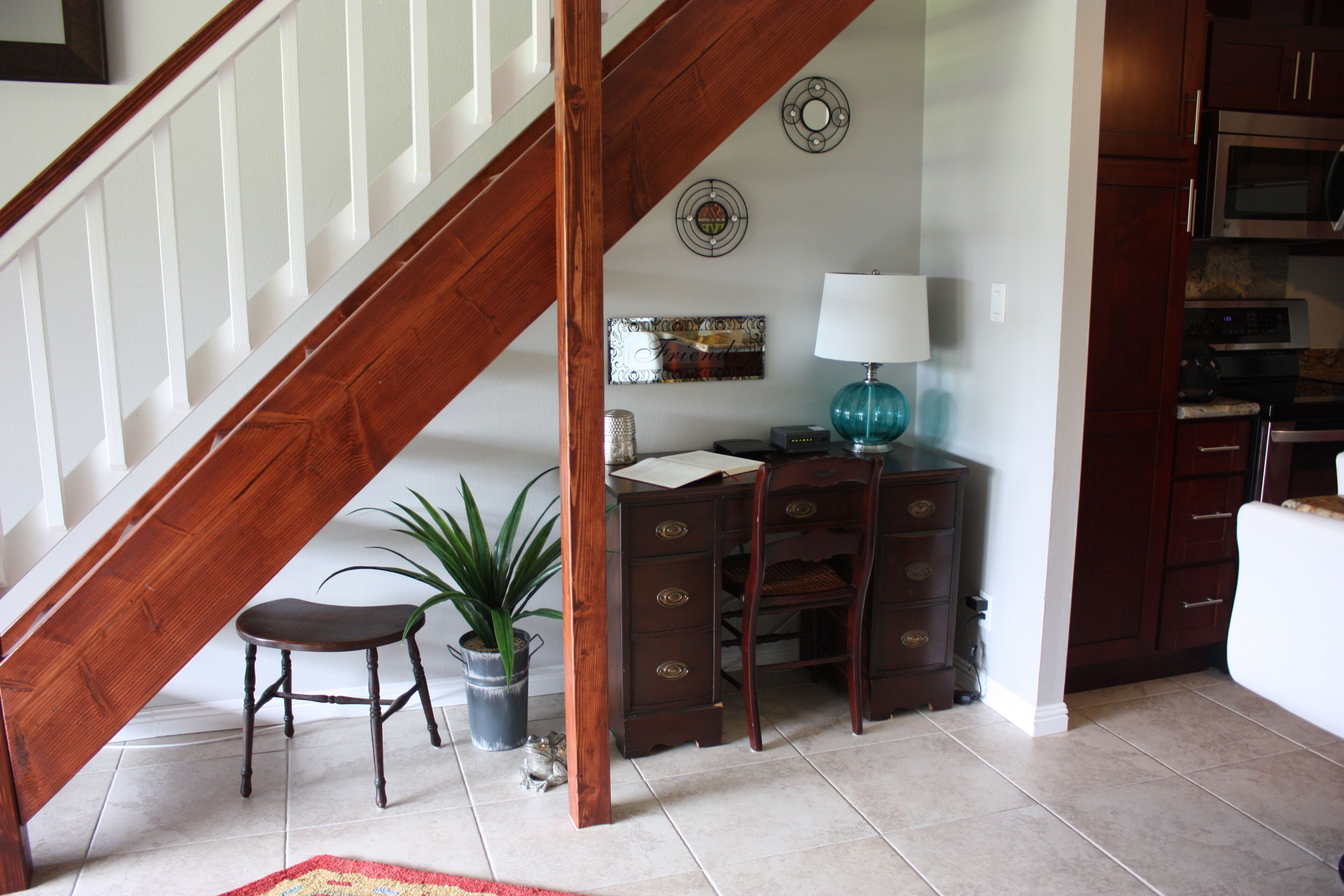 A staircase with wooden railings, a desk with drawers, a lamp, and a potted plant in a room with tiled flooring.