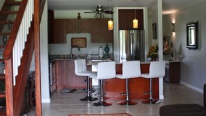 A spacious living area with a high ceiling, a kitchen with modern appliances, and a staircase leading to an upper level. - Comfortable Condo on Turtle Bay Golf Course Sleeps 5 (Kahuku)