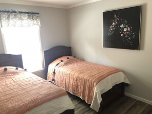 A bedroom with two single beds, a dark wooden headboard, a patterned orange and white bedspread, a black and white floral artwork on the wall, and a window with patterned curtains.