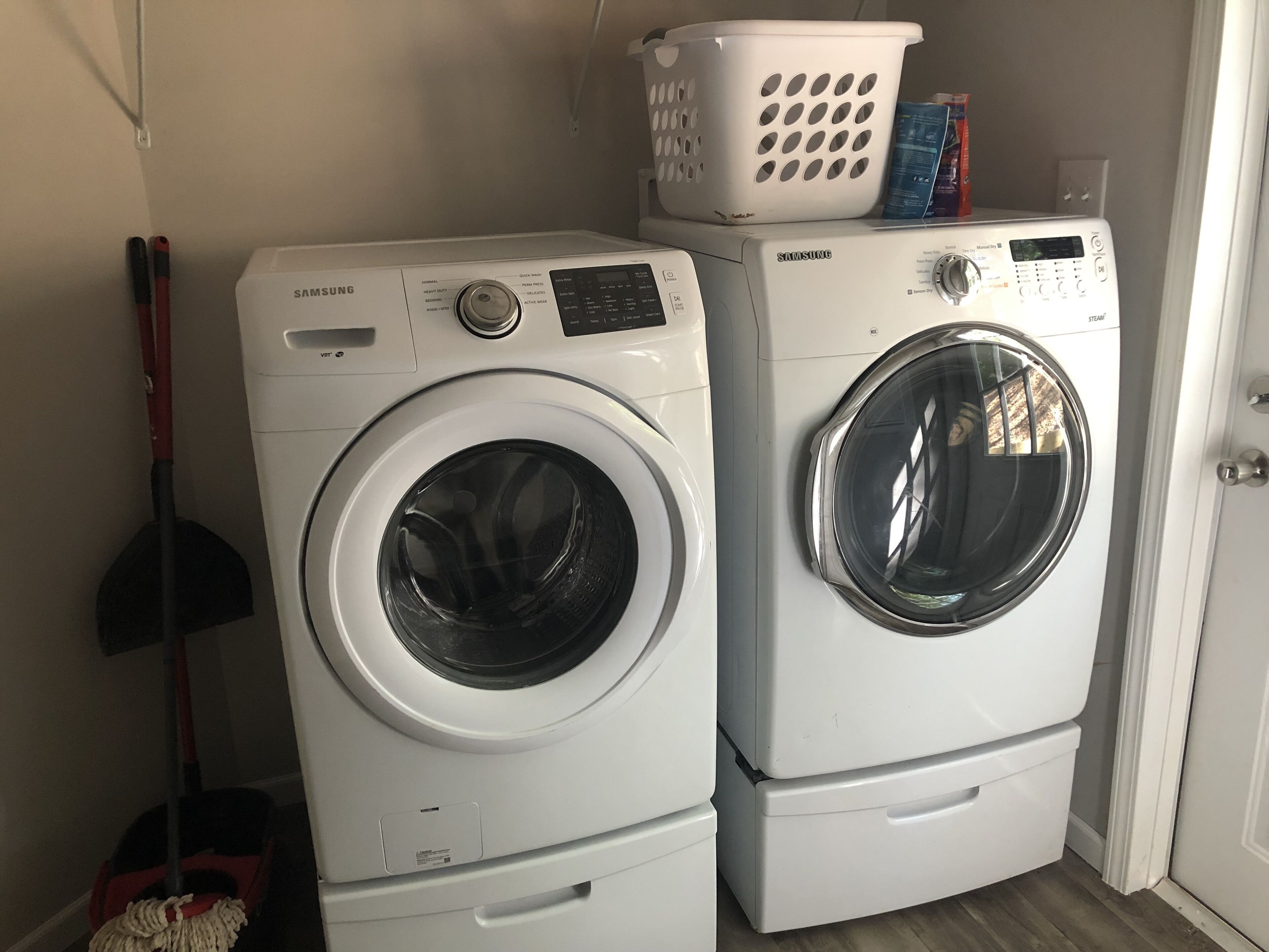 Two stacked front-loading washing machines in a laundry area with a mop and a laundry basket on top.