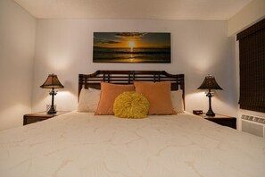A bedroom with a large bed, two bedside tables, a nightstand, a wall-mounted painting, and a window with blinds.