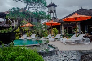 Un espace piscine agrémenté de chaises longues et de parasols, entouré de verdure et bordé d’un bâtiment traditionnel doté d’un toit en tuiles.