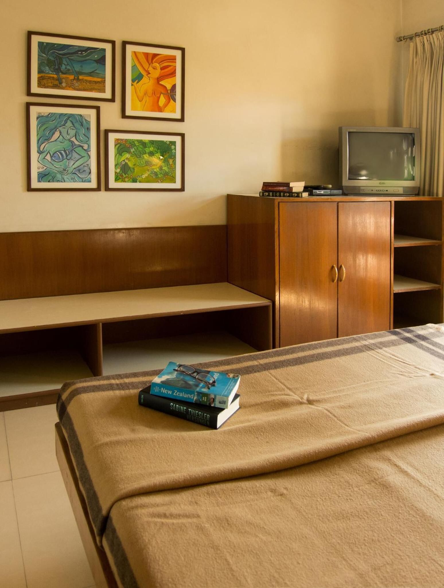 A room with a bed, a wooden bench, a TV on a cabinet, and four framed artworks on the wall.
