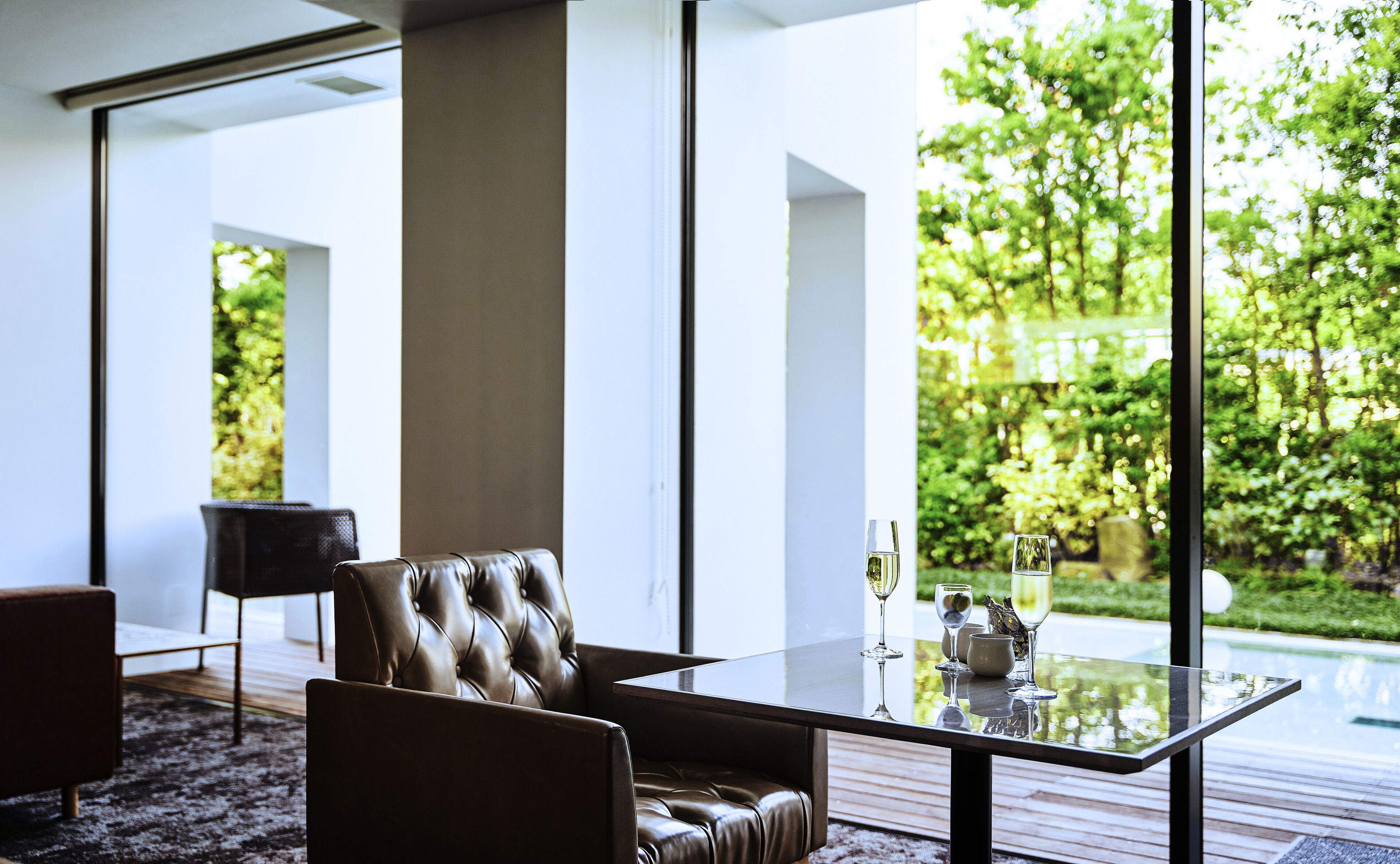 Sala de estar moderna com mesa de vidro, sofá de couro e vista para um jardim através de grandes janelas.
