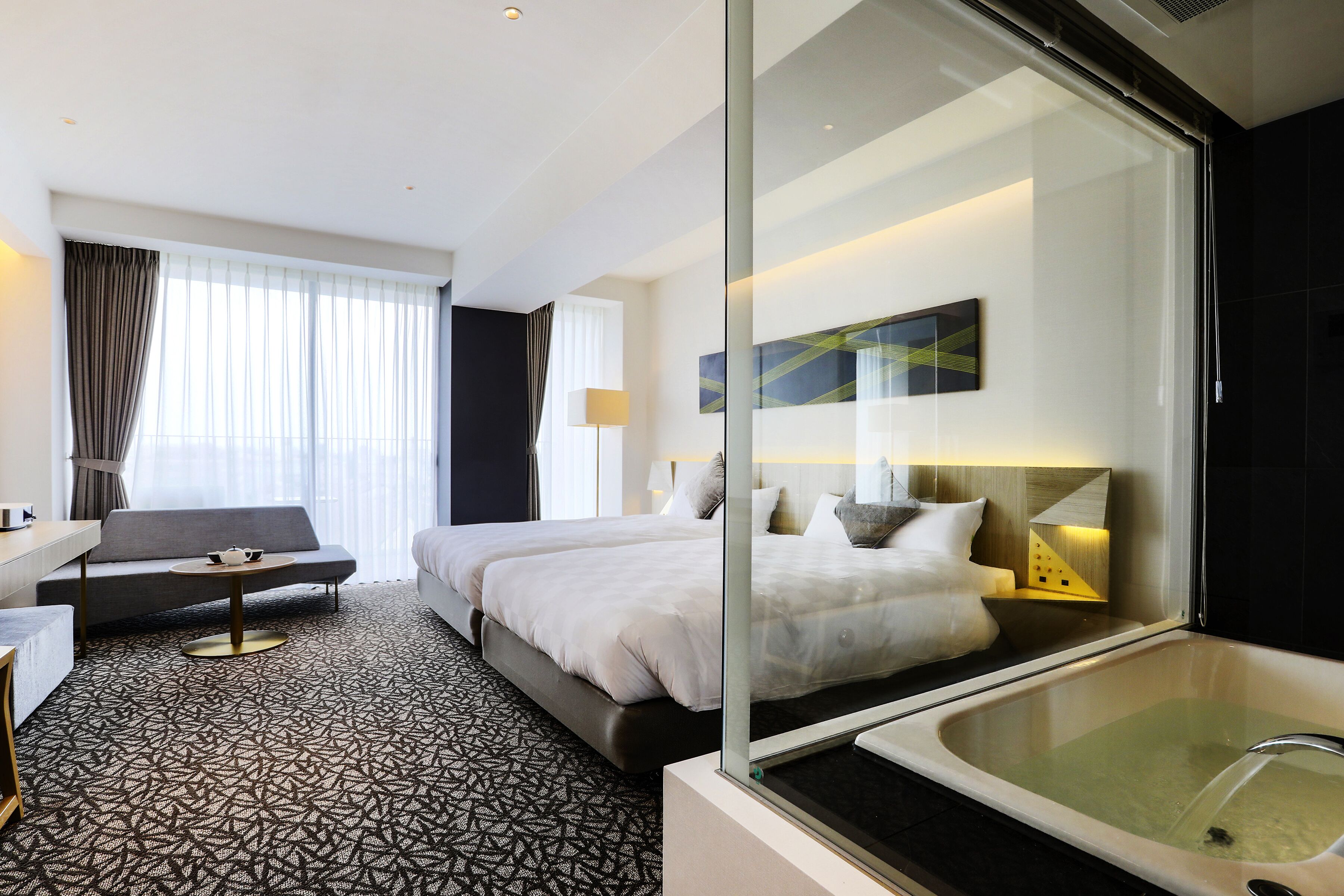 Habitación de hotel con una cama grande, un sofá, una mesita y una bañera de vidrio, además de un ventanal con cortinas.
