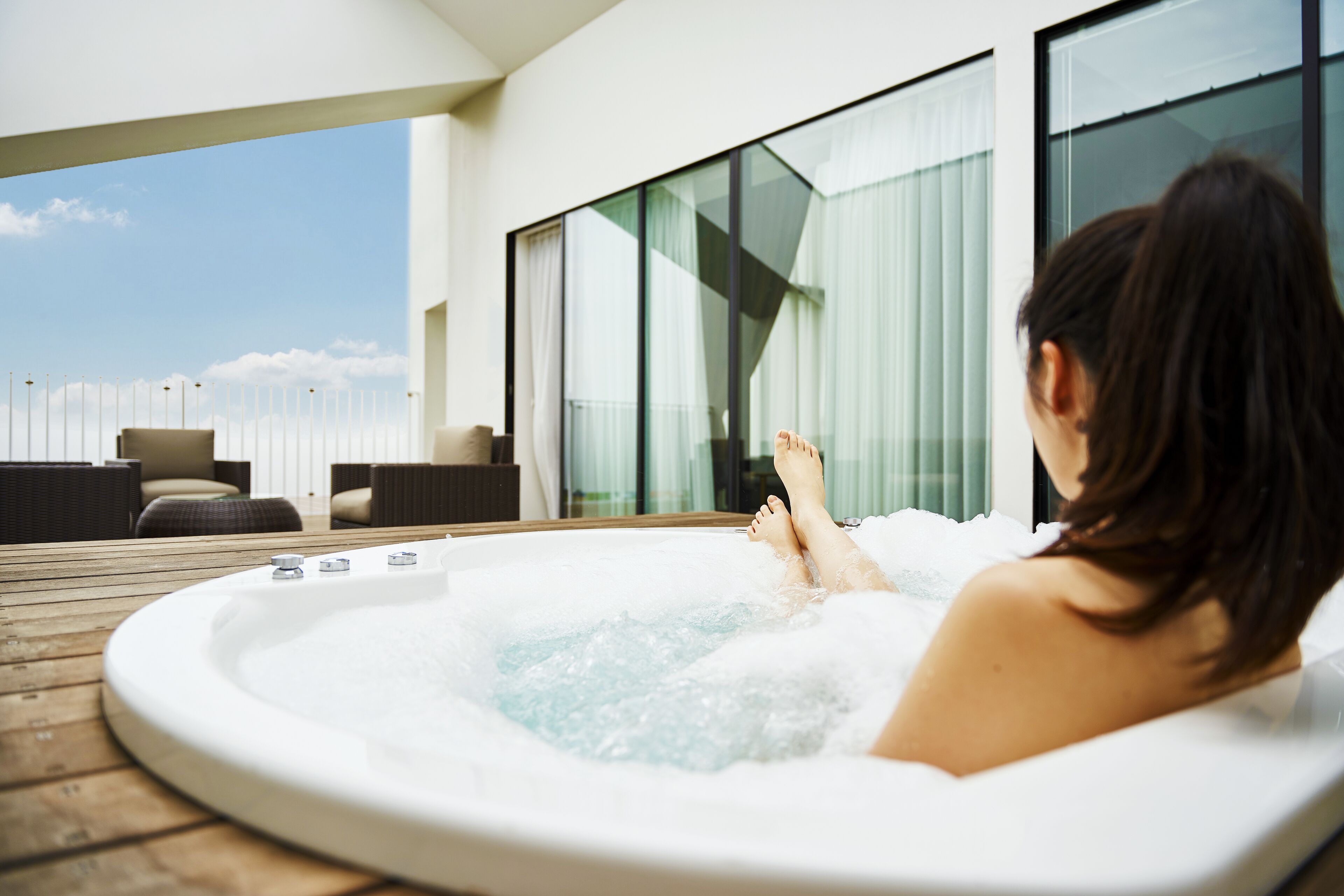 A person relaxing in a hot tub on a wooden deck with outdoor furniture and large windows.