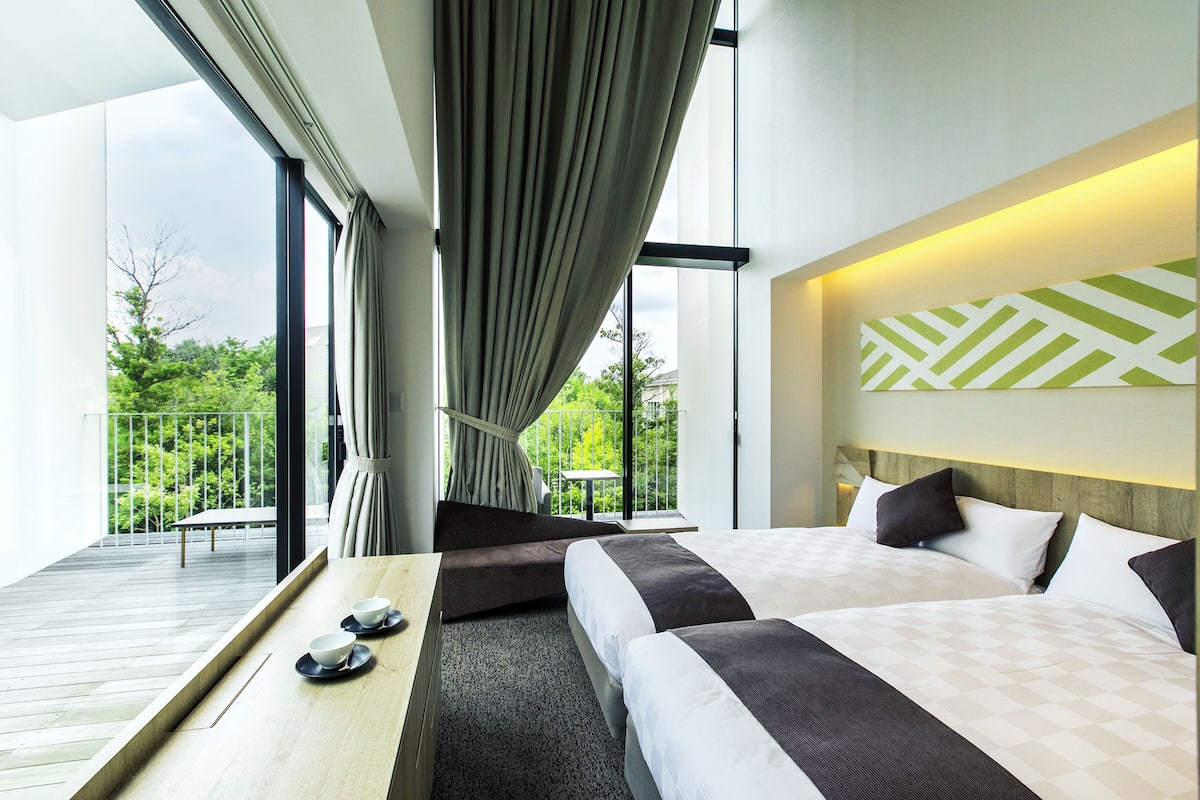 A modern hotel room with a large bed, a balcony with a table, and a view of greenery.