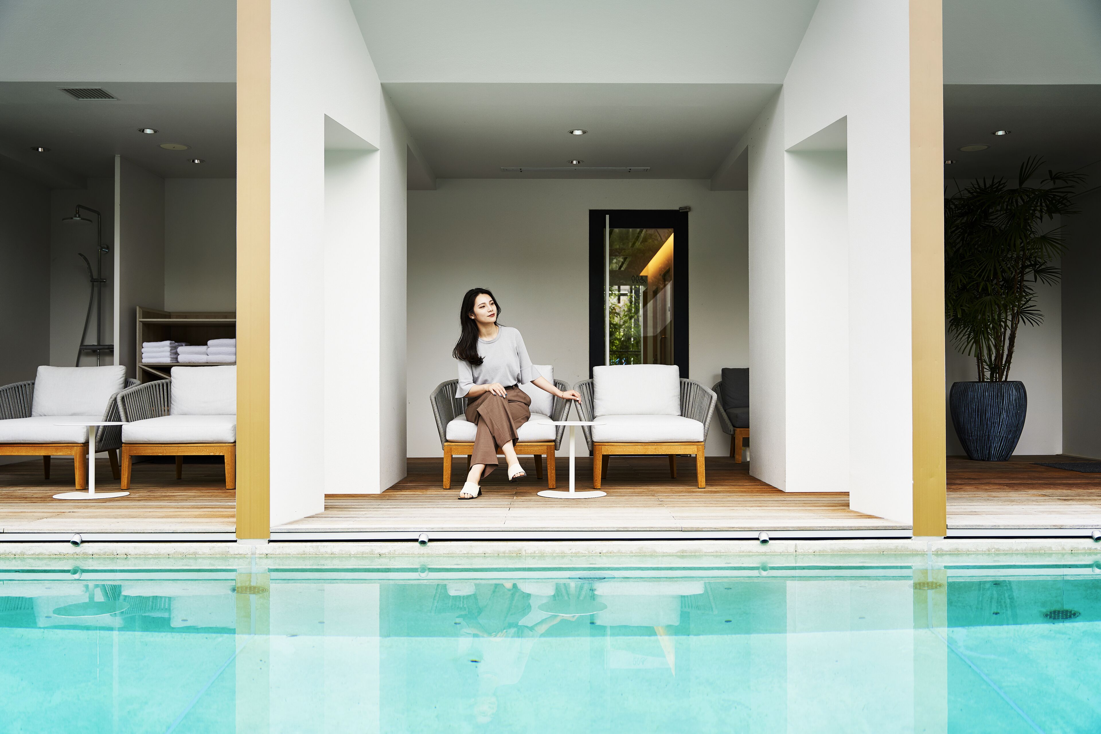A modern indoor pool area with a person sitting on a white sofa, a small table, and a potted plant.